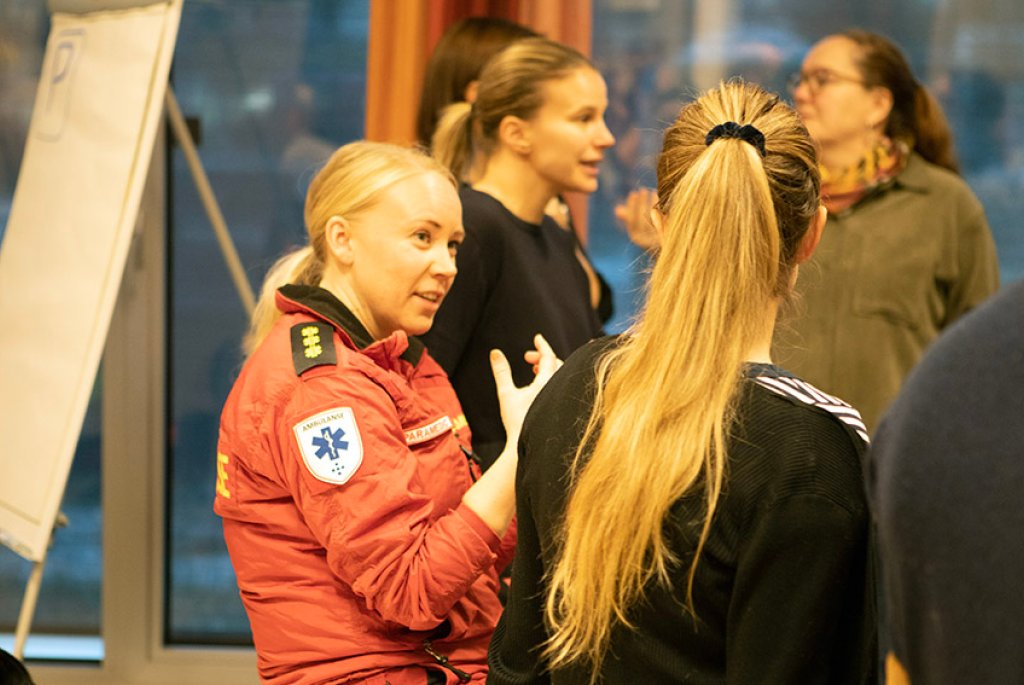 Fotografi från workshop i tjänstedesign med flera deltagare. En kvinna som jobbar inom ambulanssjukvården är i fokus och samtalar med en annan deltagare.