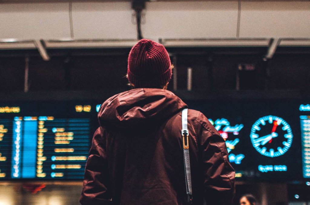 Person med jacka, mössa och väska syns bakifrån. Personen är på en flygplats och tittar på stor digital skärm som visar flygavgångar och ankomster.